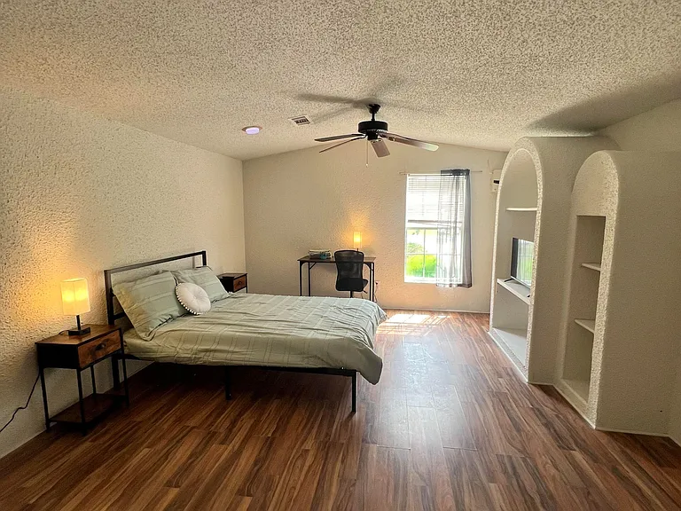 Spacious furnished bedroom with wood-look floors, ceiling fan, desk workspace, and built-in shelving for extra storage.