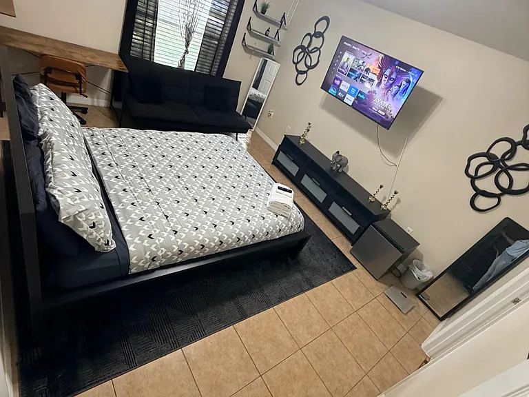 Spacious furnished bedroom with a work desk, wall-mounted TV, and seating area, plus a large window with curtains.