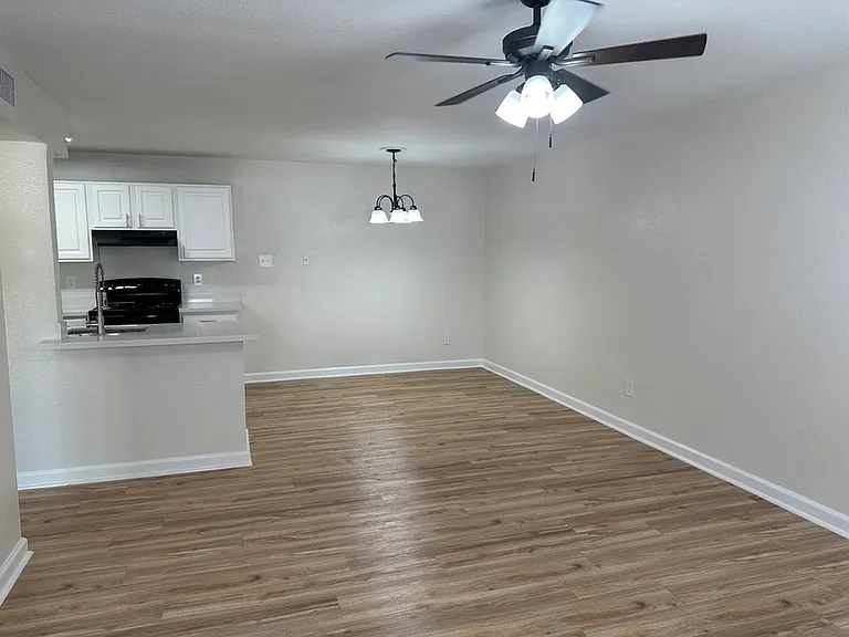 Modern open-plan living area with sleek wood-style floors, elegant ceiling fan, and a compact kitchen featuring white cabinets and black appliances. Modern open-plan living area with sleek wood-style floors, elegant ceiling fan, and a compact kitchen feat