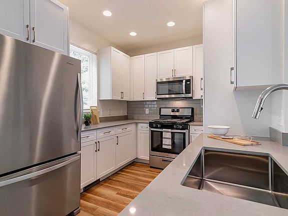 Newly renovated Finish Package III kitchen with white cabinetry, grey quartz countertops, hard surface flooring, stainless steel appliances, and tile backsplash