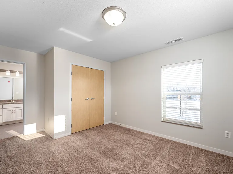 Bright bedroom with neutral carpet, large window, and closet doors, adjacent to a modern bathroom. Bright bedroom with neutral carpet, large window, and closet doors, adjacent to a modern bathroom.