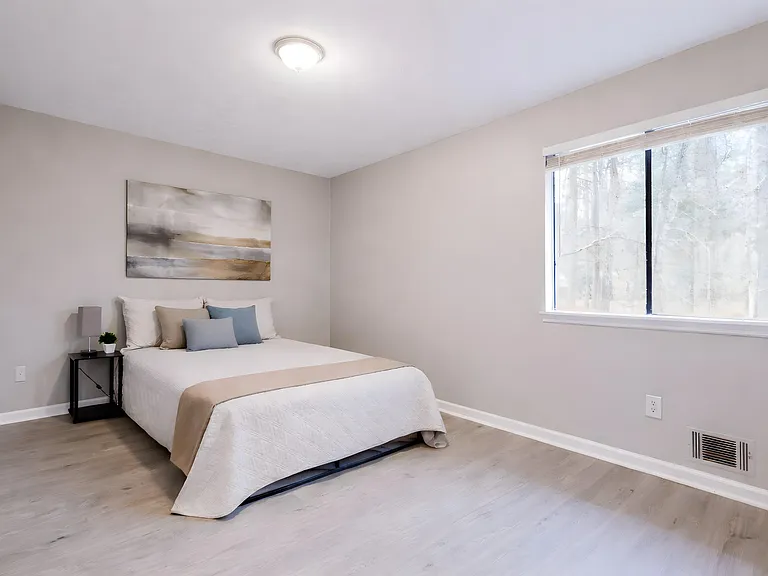 Spacious bedroom with a large window, neutral walls, and durable flooring, offering a comfortable layout for rest and work.