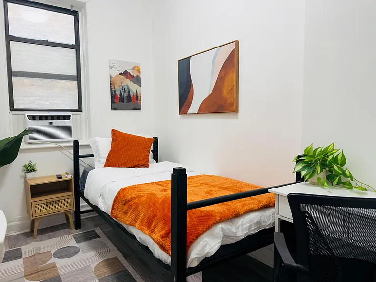 Private bedroom with a furnished bed, bedside table, and dedicated desk space, plus a window A/C unit and wall art.