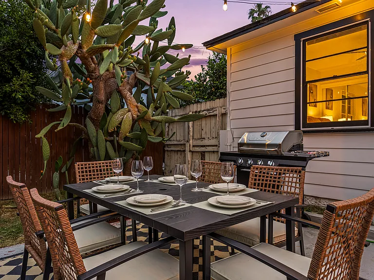 Cozy backyard retreat with cafe-style string lights, outdoor dining, a BBQ grill, and a fire pit lounge. Surrounded by palm trees and warm lighting, this inviting space is perfect for relaxing evenings and group gatherings.