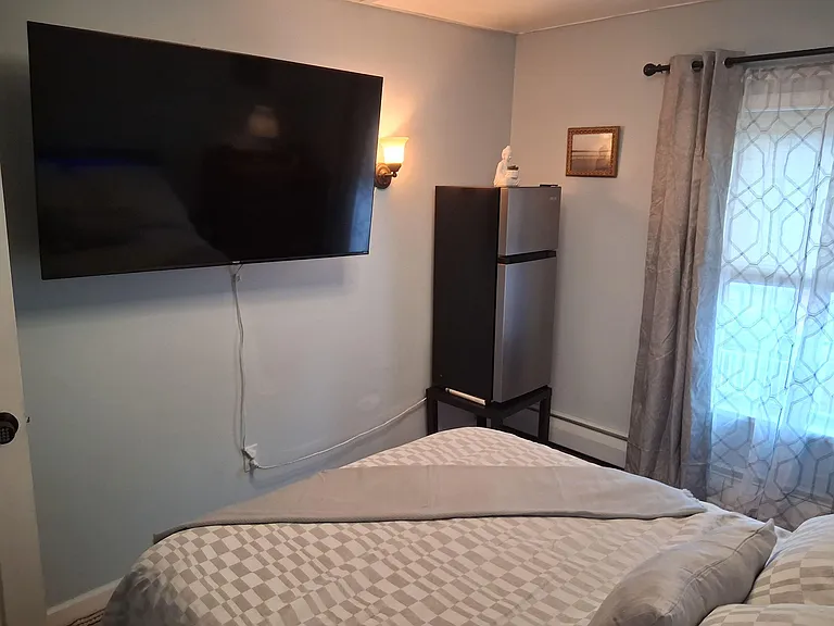 Furnished bedroom with a neatly made bed, wall-mounted TV, and window with curtains for natural light and privacy.
