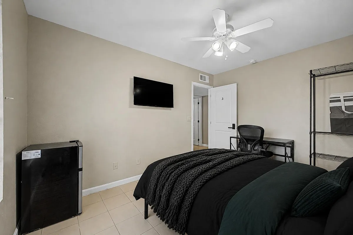 Furnished bedroom with tile flooring, ceiling fan, wall-mounted TV, mini fridge, and a dedicated desk/work area. - E 9th Ave