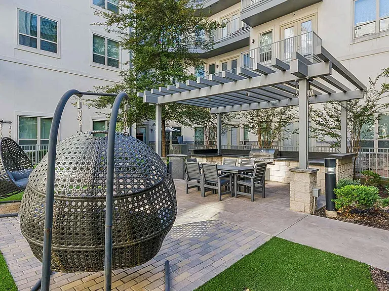 A beautifully designed courtyard in a residential complex featuring woven hanging chairs with cushions, perfect for relaxation. The space includes a pergola-covered seating area with a modern dining t