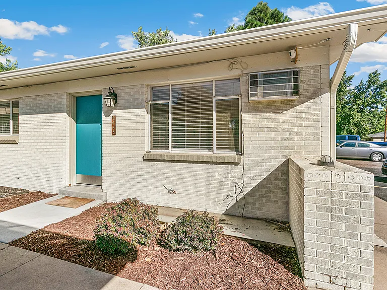 Charming apartment with a fresh teal door, surrounded by neatly landscaped greenery, offering a welcoming exterior with contemporary brickwork under a clear blue sky.
