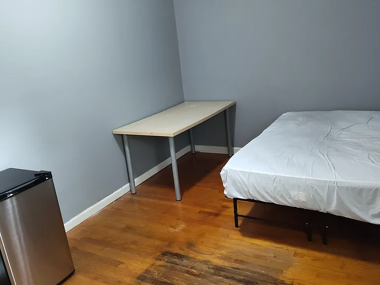 Furnished bedroom with bed, study desk, and mini fridge on hardwood floors for convenient everyday living.