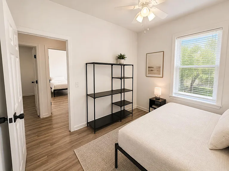 Furnished bedroom with a large window, ceiling fan, bedside table, and open shelving for convenient storage.