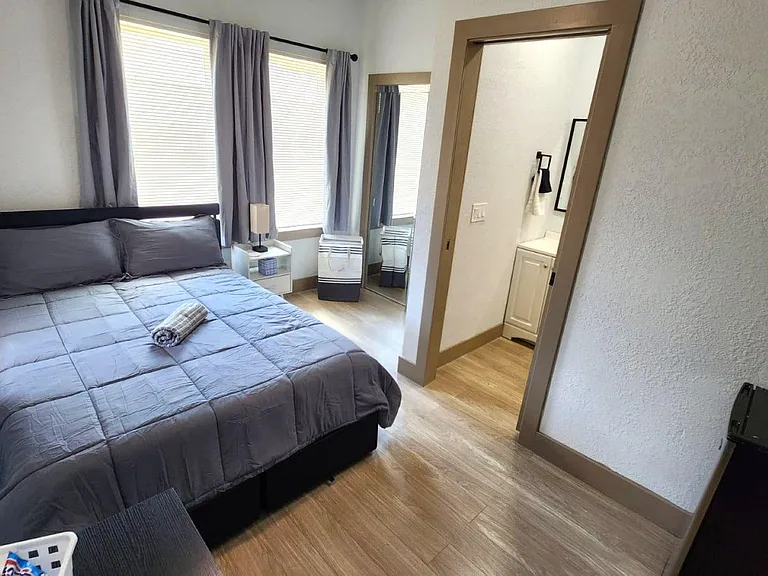 Furnished bedroom with a full bed, side table, and large windows with curtains, plus an in-room vanity area.