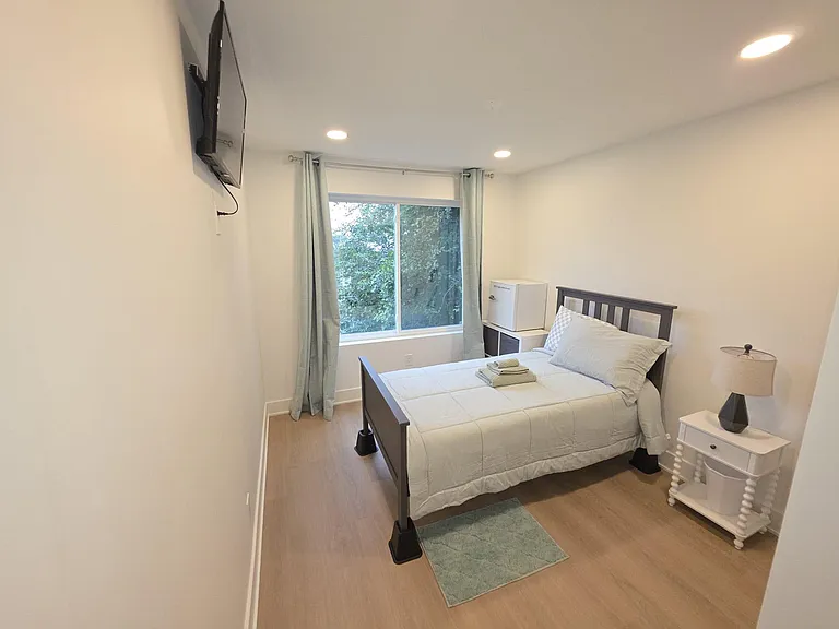 Private bedroom with a full bed, wall-mounted TV, nightstand, and a large window with curtains for natural light.