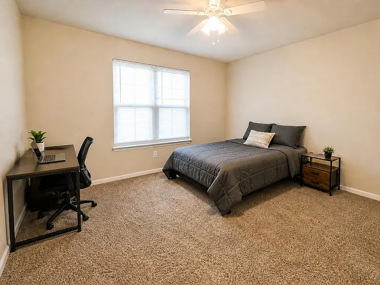Spacious bedroom with a large window, ceiling fan, and comfortable layout featuring a work desk and nightstand storage.