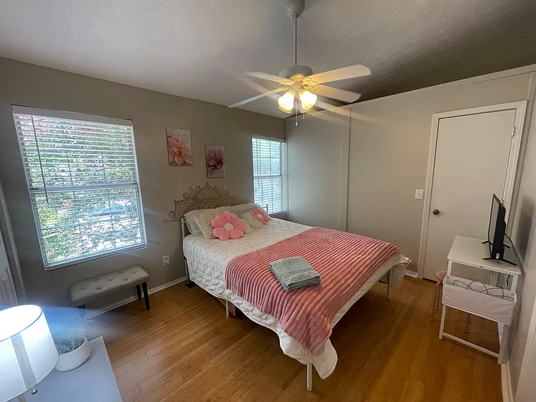 Private bedroom with hardwood floors, ceiling fan, and large windows with blinds; furnished with a bed and TV stand.