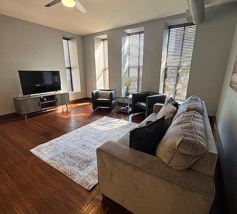 Stylish open-concept living area with a gray sofa, cozy rug, and sleek black chair. The kitchen features warm wood cabinets, stainless steel appliances, and a countertop with barstools. Dark wood floors and exposed ductwork add an industrial-modern vibe.