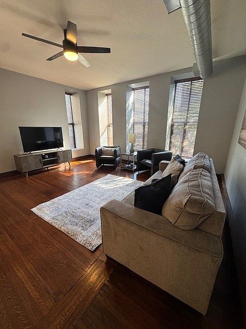 Stylish open-concept living area with a gray sofa, cozy rug, and sleek black chair. The kitchen features warm wood cabinets, stainless steel appliances, and a countertop with barstools. Dark wood floors and exposed ductwork add an industrial-modern vibe.