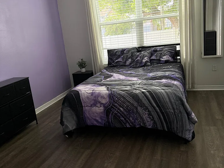 Comfortable bedroom with a large window, ceiling fan, and wood-style flooring, plus space for a nightstand and dresser.