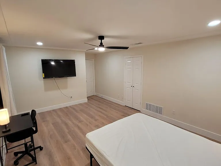 Spacious furnished bedroom with ceiling fan, wall-mounted TV, dedicated desk area, and double-door closet for storage.