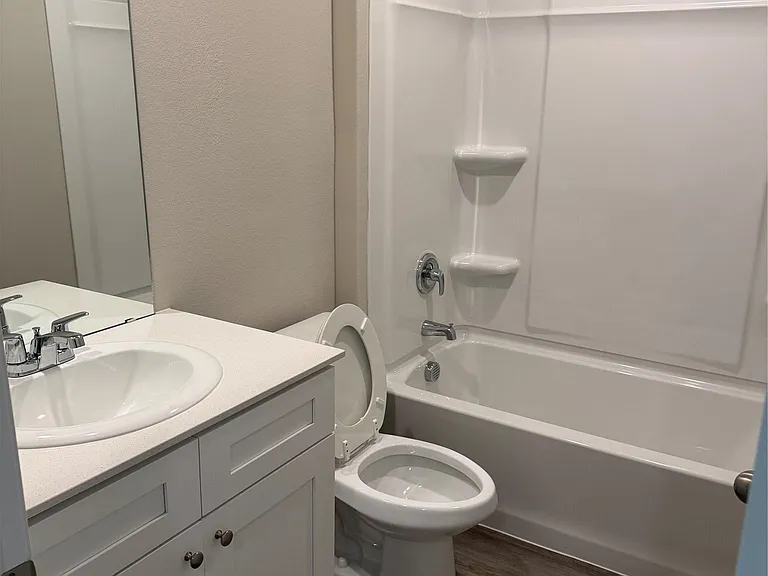 Bathroom featuring a white vanity with an integrated sink, a full-width wall mirror, a toilet, a white shower-over-tub unit with built-in shelving, and wood-finish flooring