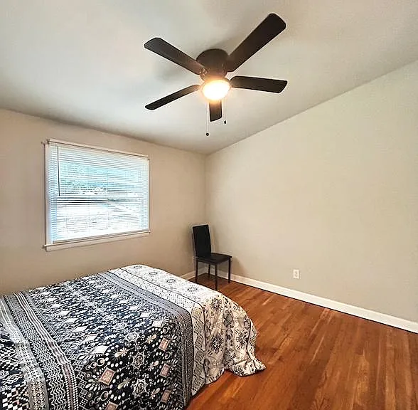 Bedroom with hardwood floors, a ceiling fan with light, and a window with blinds; ample space for a full bed and seating.