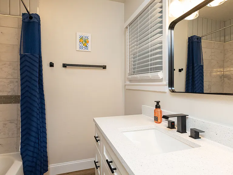 Bright, clean bathroom featuring a modern vanity with black fixtures and a fully tiled tub/shower combo. Marble-style walls, a bold blue curtain, and sleek accents create a fresh, polished space for getting ready or unwinding.