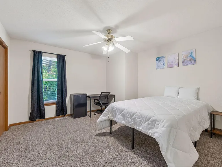 Spacious bedroom with a large window, ceiling fan, and soft carpeting, plus a desk area and mini fridge for convenience.