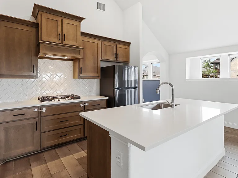 Spacious kitchen featuring an island with a stainless steel sink and gooseneck faucet, light-toned countertops, wood-finish cabinetry, a herringbone tile backsplash, and wood-finish flooring