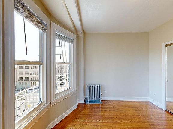 Bright room with hardwood floors, two large windows, a wall-mounted radiator, and a mirrored closet door.