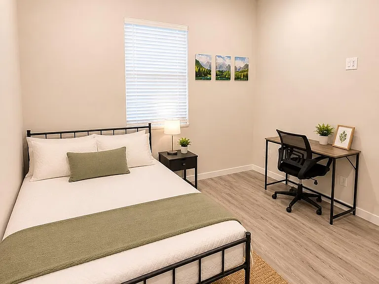 Furnished bedroom with a full bed, nightstand, and dedicated desk area, plus a ceiling fan and window with blinds.