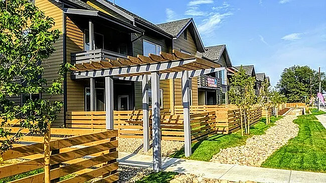 Charming multi-story apartments with wooden accents, lush landscaping, and inviting pergola entryway under a clear blue sky.
