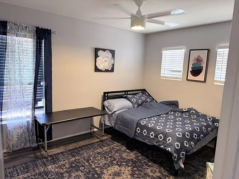 Furnished bedroom with a twin bed, desk workspace, ceiling fan, and multiple windows with blinds for privacy.