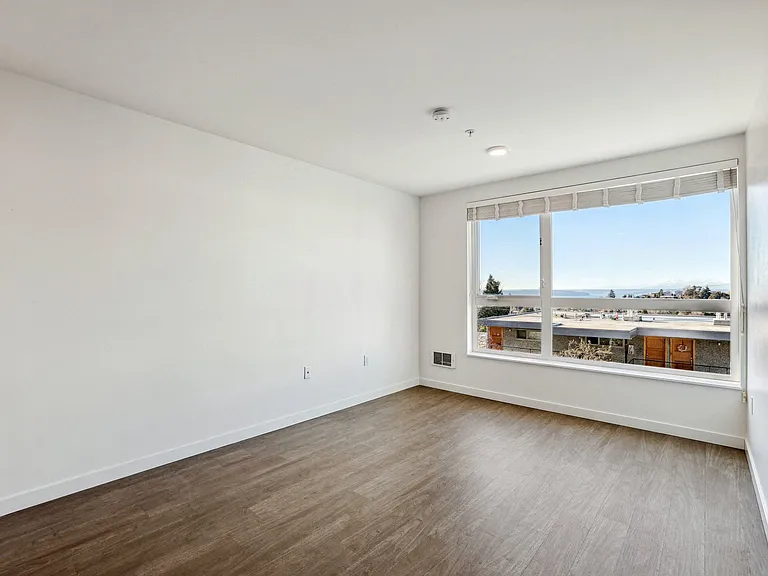 Spacious, unfurnished room at 4400 Alaska in Seattle, WA, featuring bright natural light, wood-look flooring, and stunning views of the water and mountains.