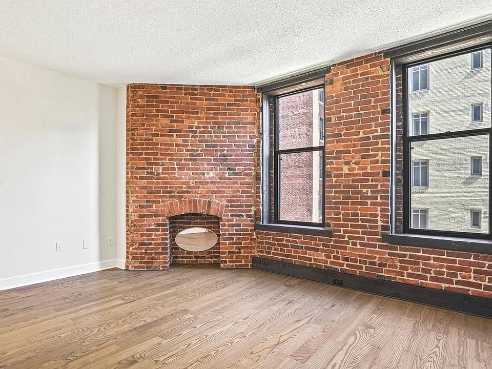 Living room with hardwood floors, exposed brick wall, large windows, high ceilings, and decorative fireplace. Note: Mirror in decorative fireplace is not included.