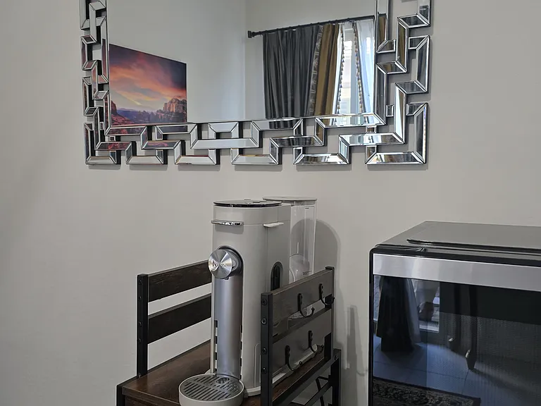 The lounge features a stylish coffee station with espresso machine and coffee pods, plus a sleek silver fridge and microwave combo nearby. Coordinated metallic finishes and thoughtful details create a welcoming, well-equipped space.