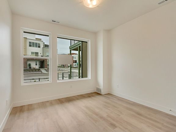 A bright, empty room with light wood flooring, white walls, and two large windows that overlook a mo