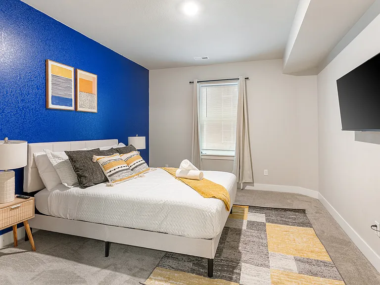 Bright and inviting bedroom with a royal blue accent wall and modern decor. Features a comfortable queen bed with white linens, gray and yellow pillows, bedside tables with lamps, a colorful geometric rug a window with curtains, and a wall-mounted TV