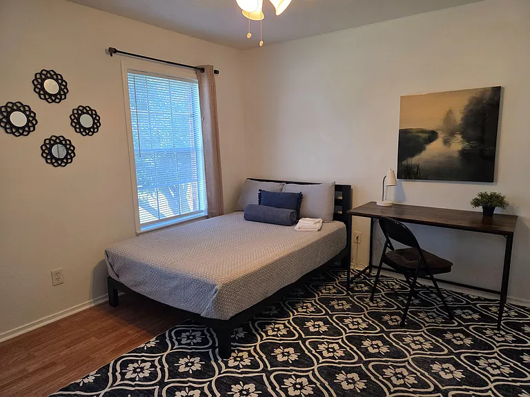 Furnished bedroom with a comfortable bed, work desk, and a large window with blinds for natural light and privacy.