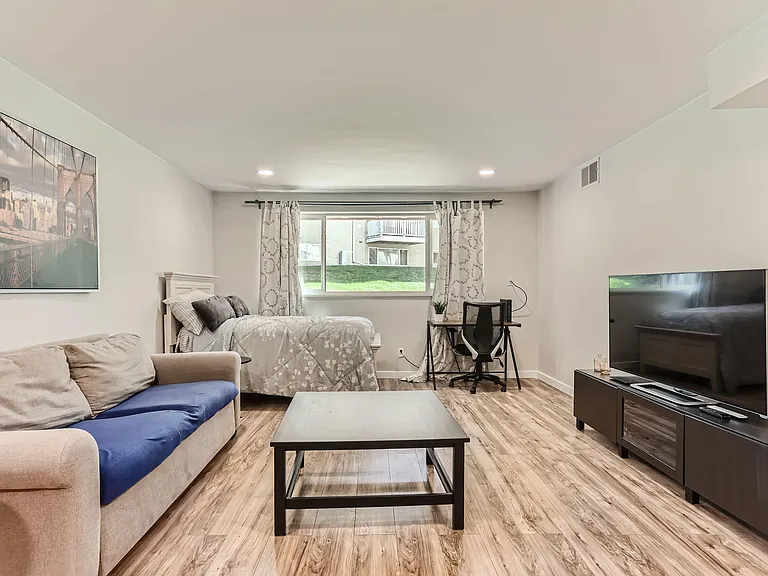 Spacious studio-style room featuring a comfortable bed, lounge seating, dedicated desk area, and large window with curtains.