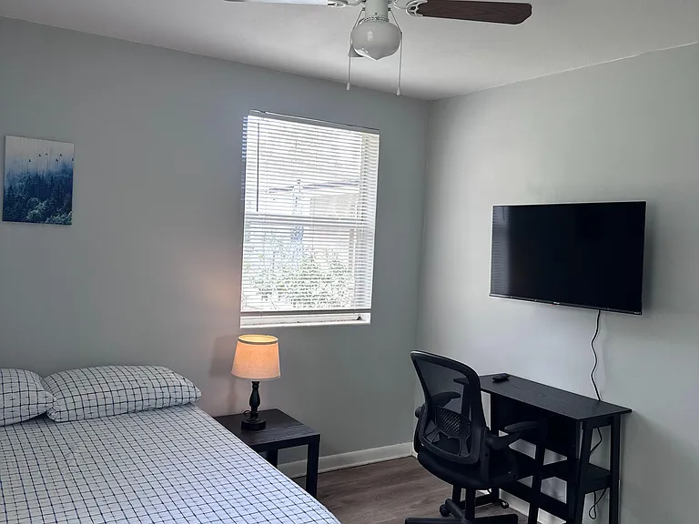 Furnished bedroom with a ceiling fan, window blinds, bedside table with lamp, and a dedicated desk/workspace area.