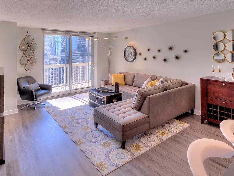 Looking into a living room with a sliding glass window leading to a balcony on the far end. A sitting area is on the right with a large couch and table.