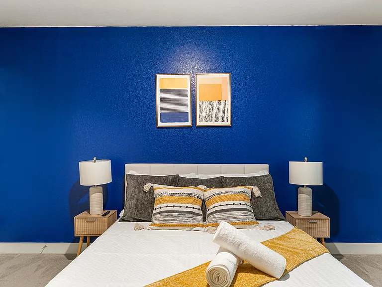 Cozy bedroom featuring a bold royal blue accent wall with two modern art pieces. The bed is dressed in white linens with gray and yellow decorative pillows, complemented by matching bedside tables and lamps, creating a stylish and inviting atmosphere