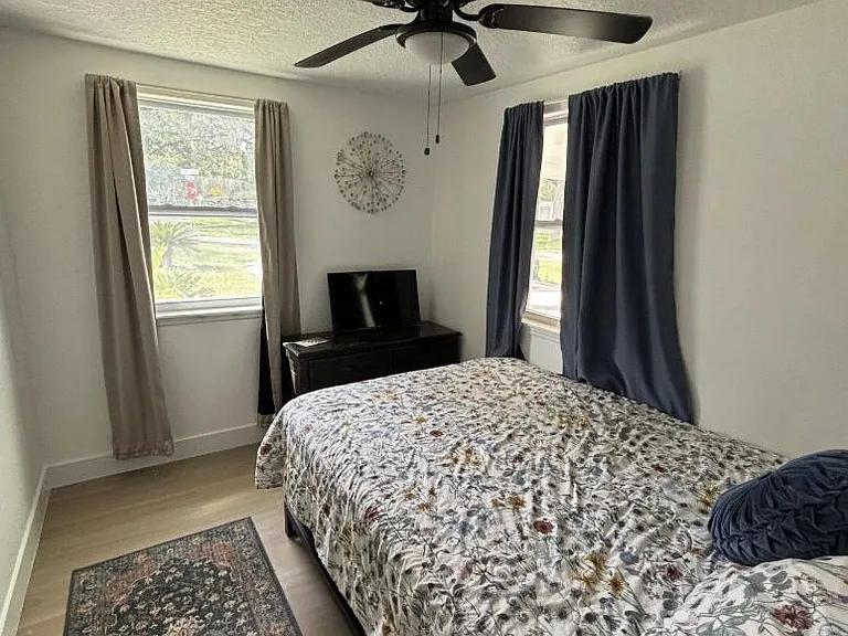 Furnished bedroom with a ceiling fan, two windows with curtains, and space for a dresser and bedside essentials.