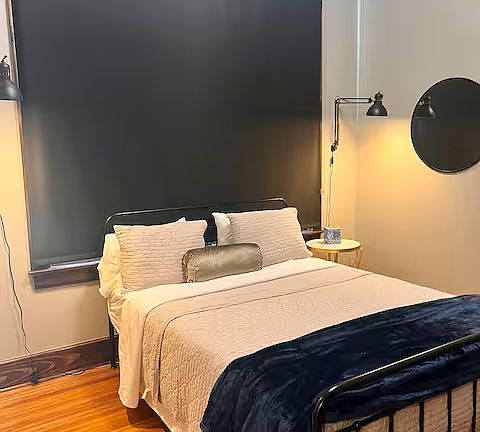 The bedroom is a cozy retreat with a plush queen bed, soft linens, and warm pillows. Blackout shades ensure total darkness for restful sleep. A bedside lamp casts a gentle glow, and a nightstand holds essentials. Compact yet inviting.