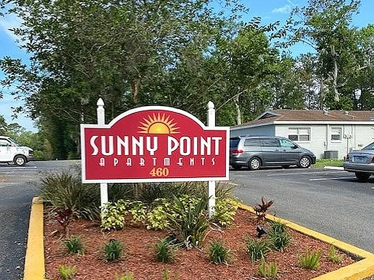 Inviting entrance to Sunny Point Apartments with vibrant landscaping and ample parking space.