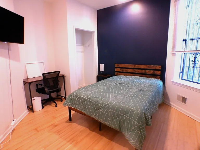 Furnished bedroom with hardwood floors, a work desk, wall-mounted TV, and an open closet plus a window with blinds.