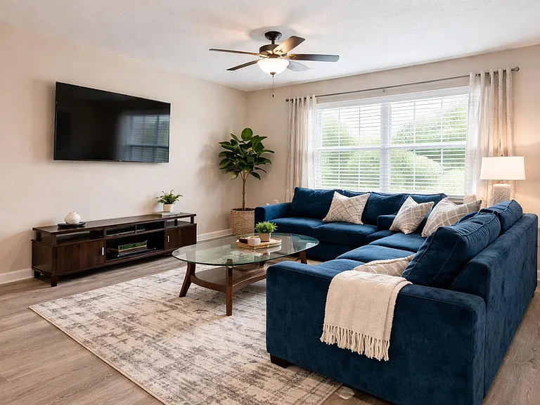 Spacious living room with a large sectional, wall-mounted TV, and ceiling fan, plus a wide window with blinds and curtains.