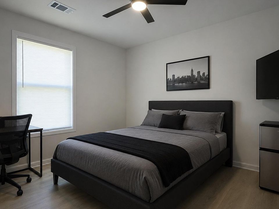 Furnished bedroom with a work desk, ceiling fan, wall-mounted TV, and mini fridge, plus a large window with blinds.