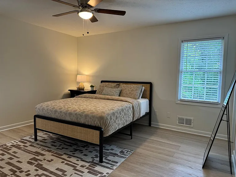 Furnished bedroom with a full-size bed, ceiling fan, and a window with blinds for natural light and privacy.