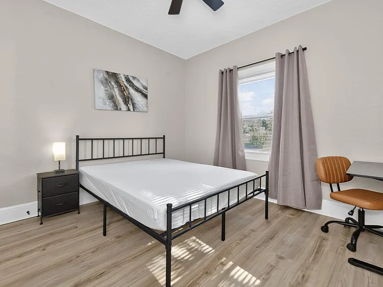 Private bedroom with a full bed, nightstand lighting, and a dedicated desk area, plus a window with curtains.