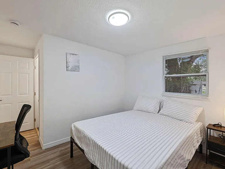 Furnished bedroom with a full bed, work desk, and nightstand, plus a window with blinds for natural light.
